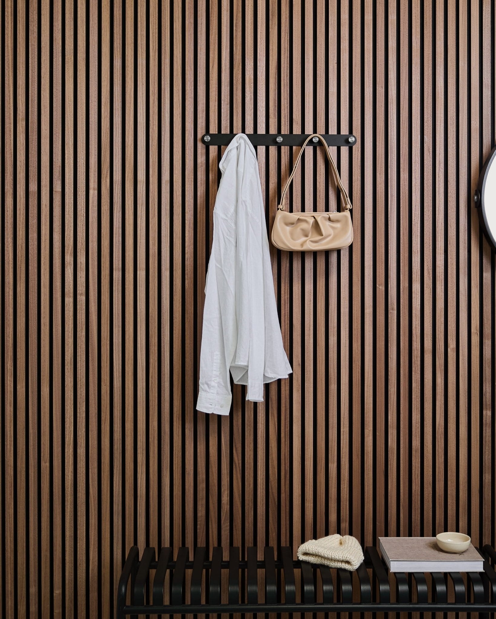 Een wit overhemd en beige handtas hangen aan WoodUpps Norr Coat Hook Rack boven een bankje, geplaatst voor een houten Akupanel lattenwandpaneel met decoratieve items.